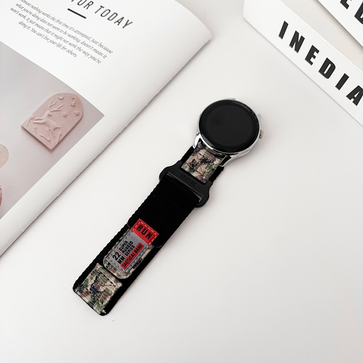 Smartwatch con cinturino tattico in nylon nero su una rivista, con inserti mimetici e un'etichetta rossa con la scritta 'RUN'.