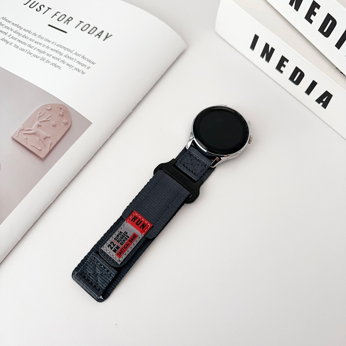 Eine graue Uhr mit einem Nylonarmband befindet sich auf einer weißen Oberfläche mit Büchern und einer Statue.