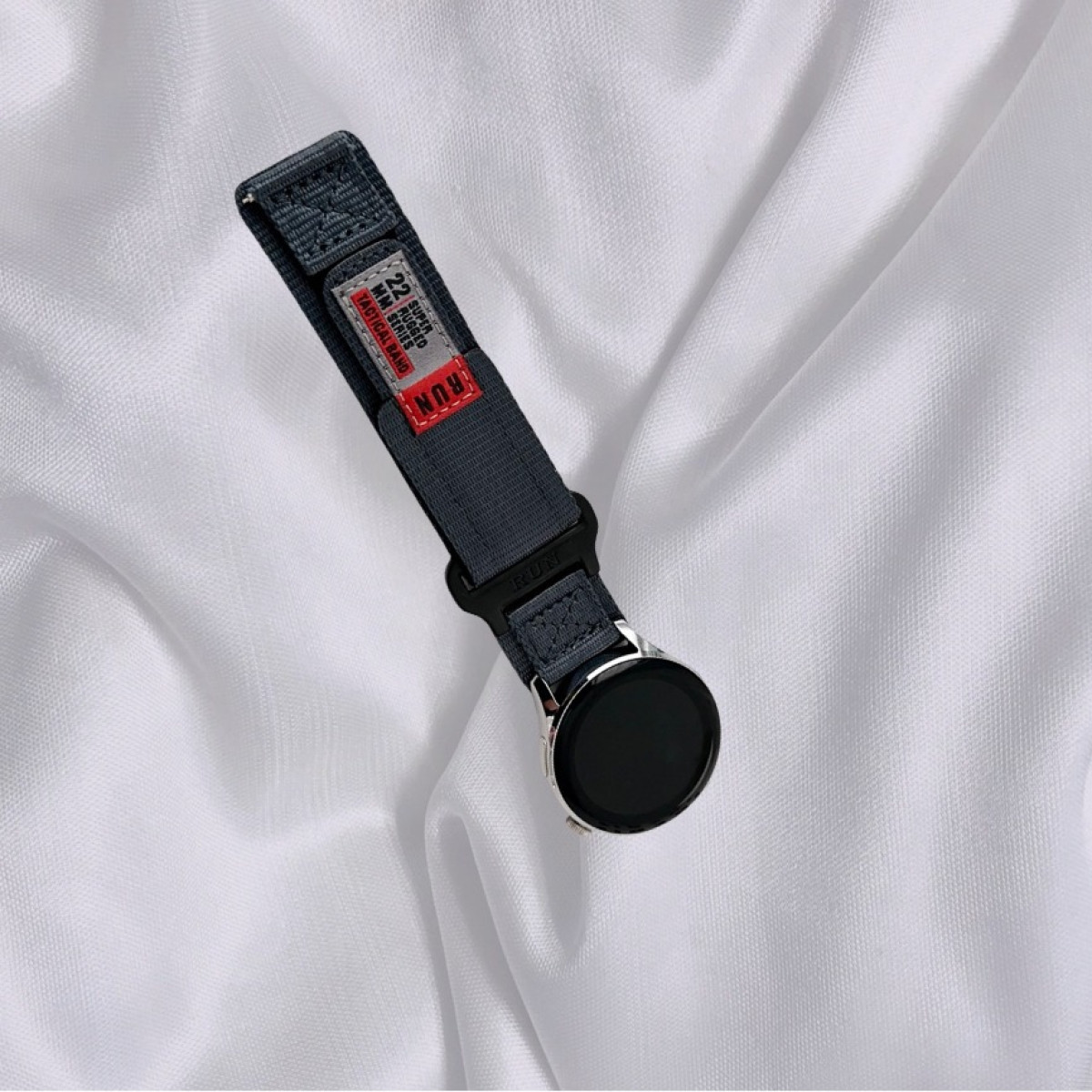 Eine graue Uhr mit einem Nylonarmband wird auf einem strukturierten weißen Stoffhintergrund angezeigt.
