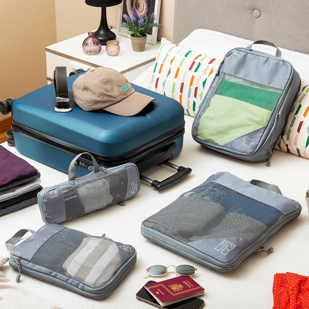 Vier graue Packwürfel auf einem Bett mit einem blauen Koffer, Hut und anderen Reiseartikeln.