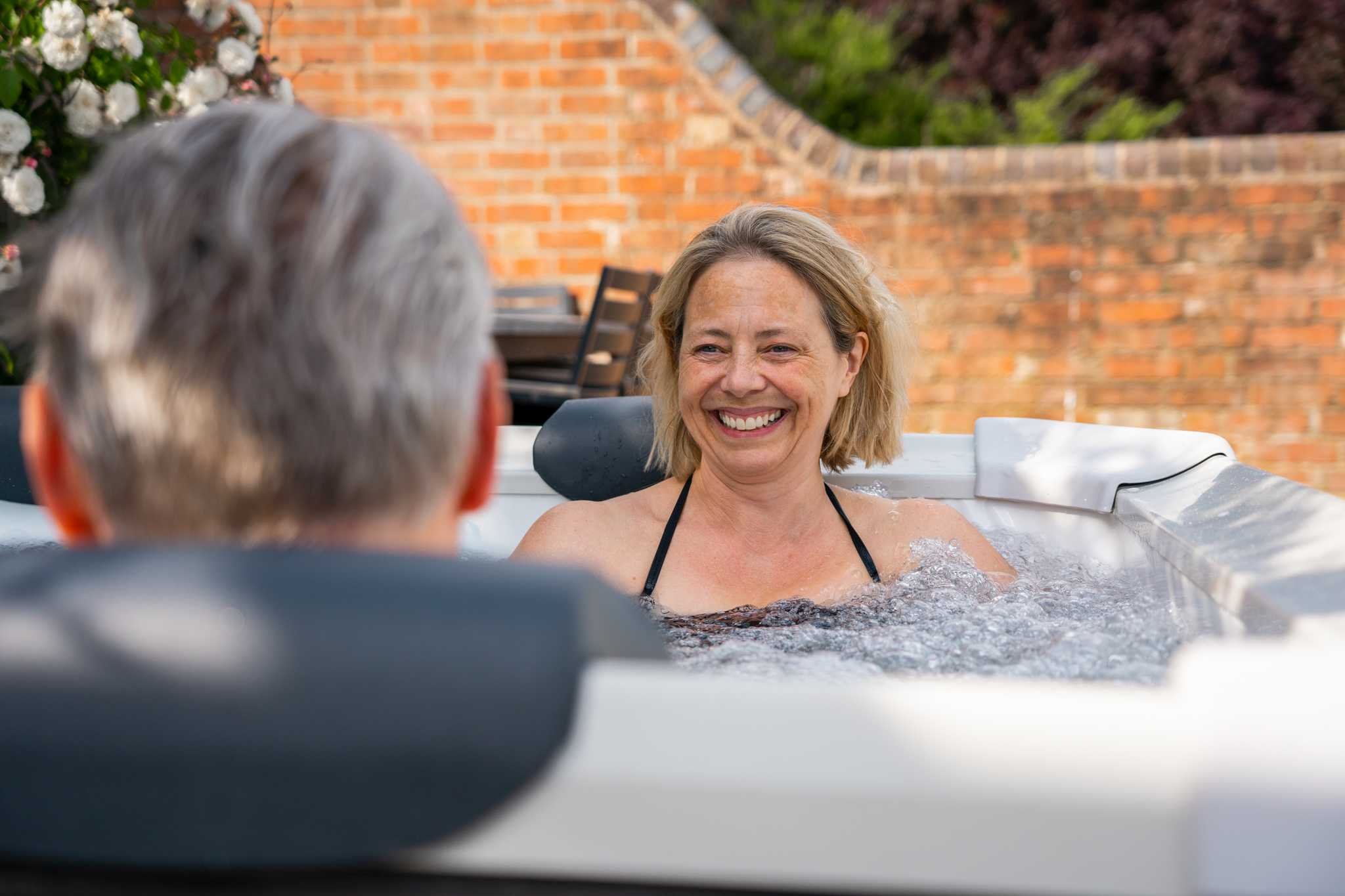 Kobieta uśmiecha się w bulgoczącej wannie z hydromasażem z mężczyzną, za nią ceglana ściana.