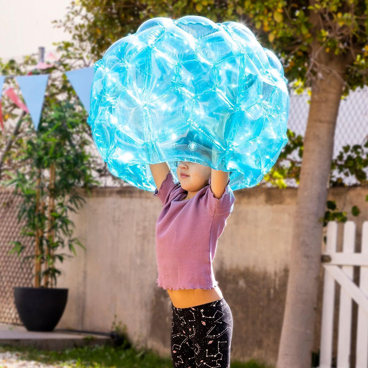 Un niño con una camisa morada sostiene una gran bola inflable azul por encima de la cabeza en un entorno soleado al aire libre.