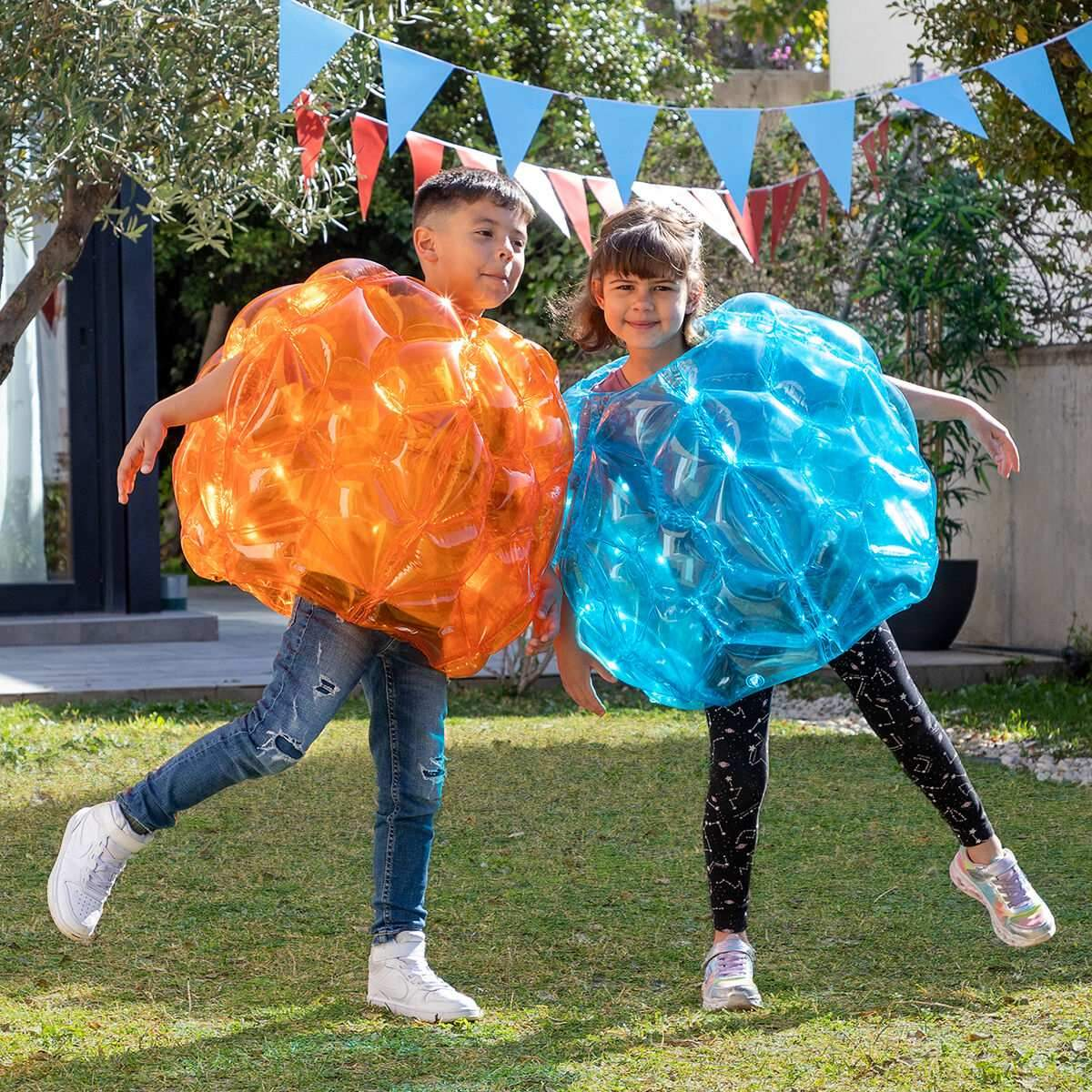 Dos niños en pelotas inflables posan al aire libre sobre un césped bajo pancartas festivas.