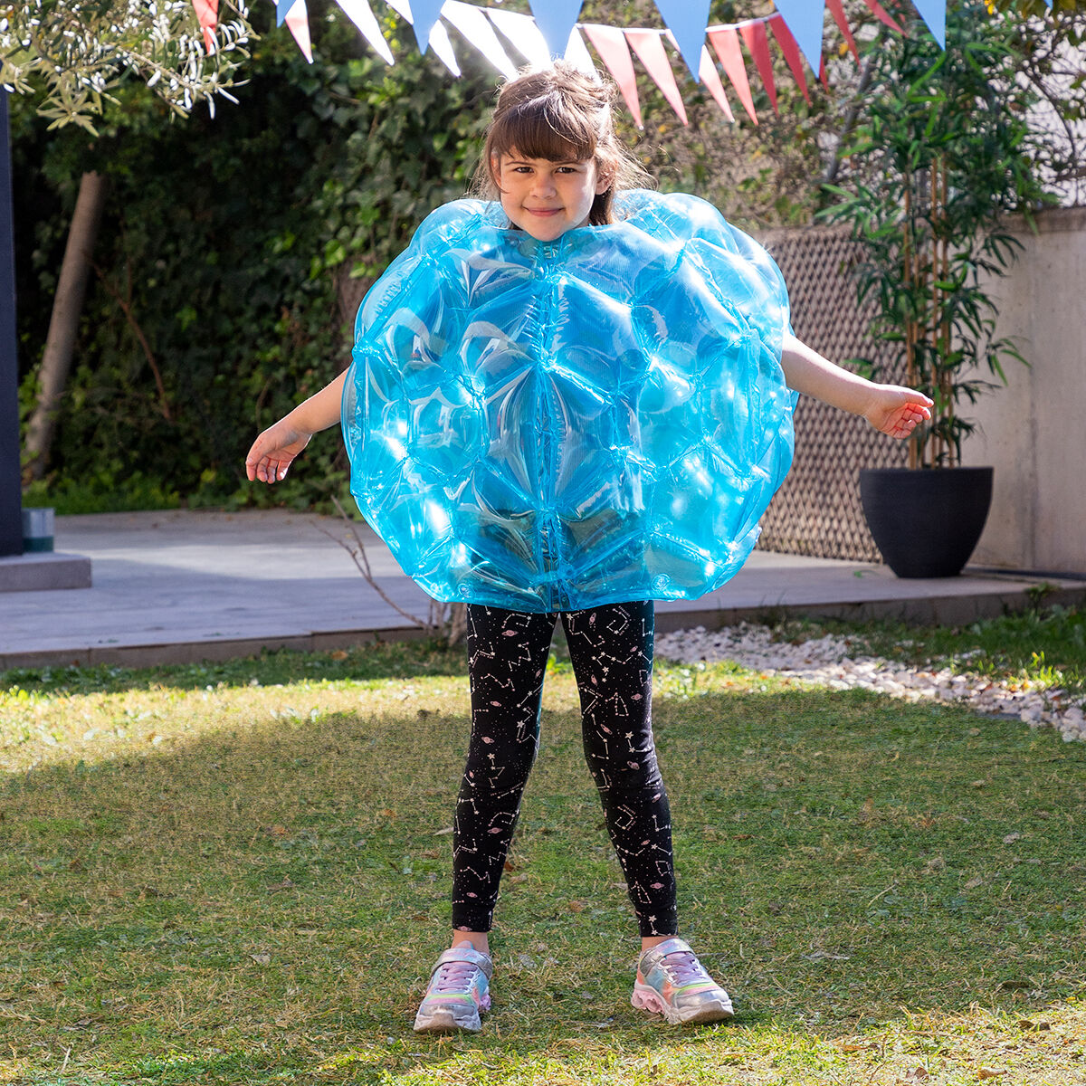 Una niña está de pie en un césped con un traje de bola azul inflable grande, sonriendo a la cámara.