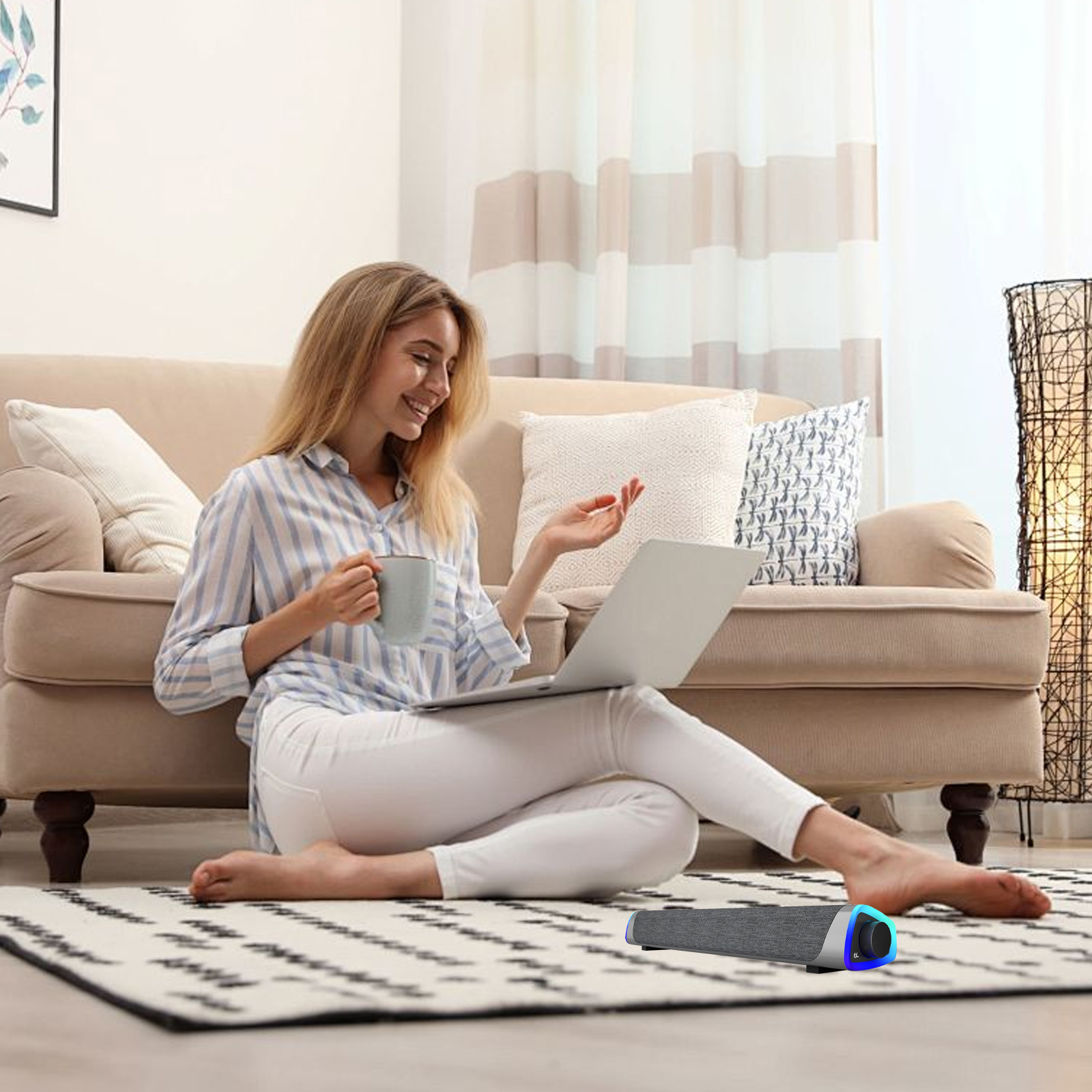 Eine Frau sitzt auf dem Boden und benutzt einen Laptop, mit einer grauen Soundbar in der Nähe und einem Sofa und einer Lampe im Hintergrund.