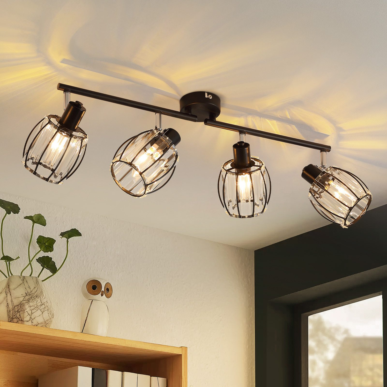 Un lampadario a gabbia a quattro lampadine illumina una stanza con una pianta, una statuetta di gufo e una mensola di legno.