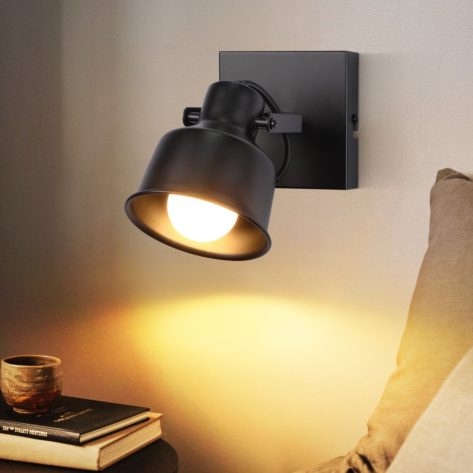 Un faretto nero è montato su una parete, illuminando un comodino con un libro e una tazza. La luce è calda e concentrata.