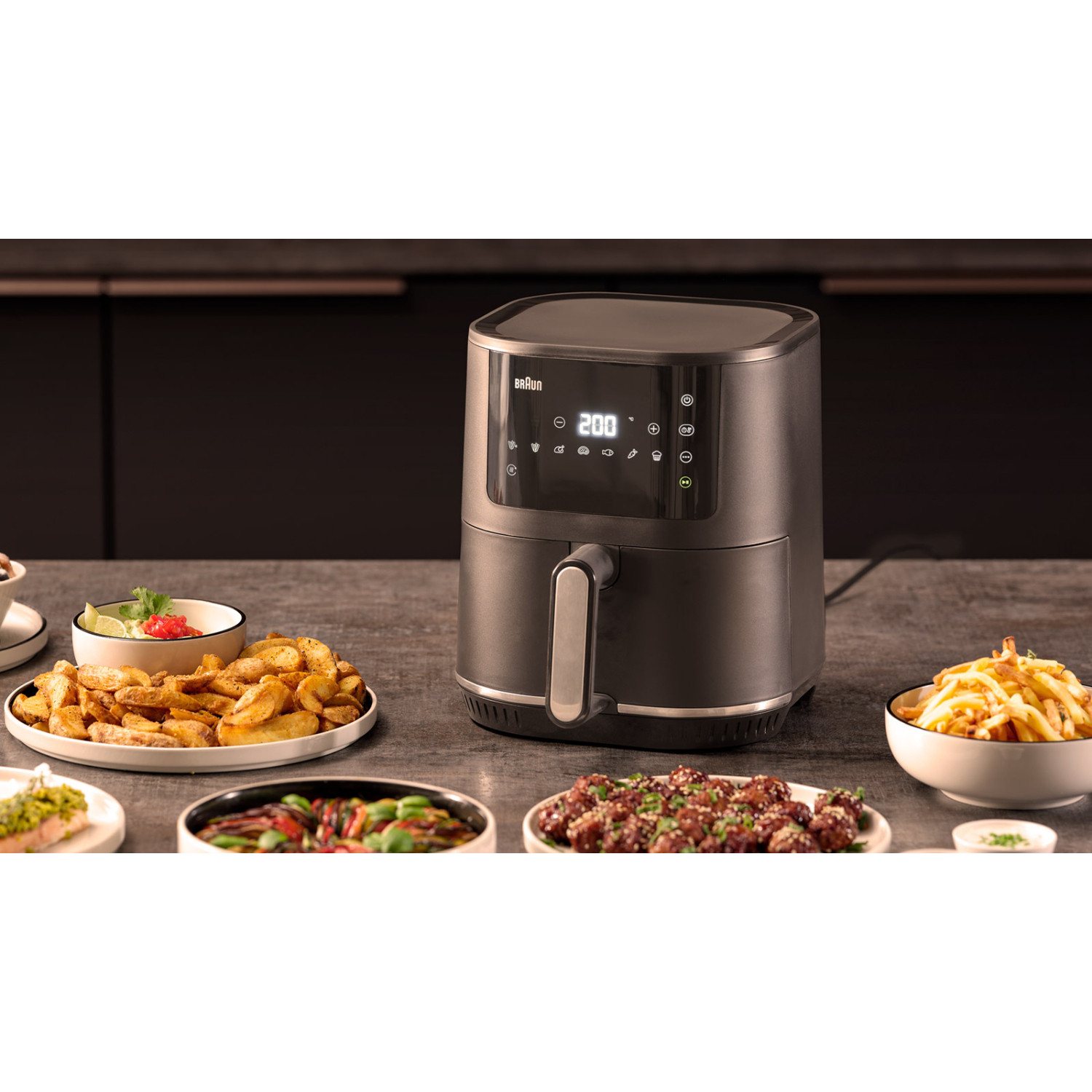 Eine schwarze Braun Fritteuse auf einer Arbeitsplatte, umgeben von Tellern mit gekochtem Essen vor einem dunklen Hintergrund.