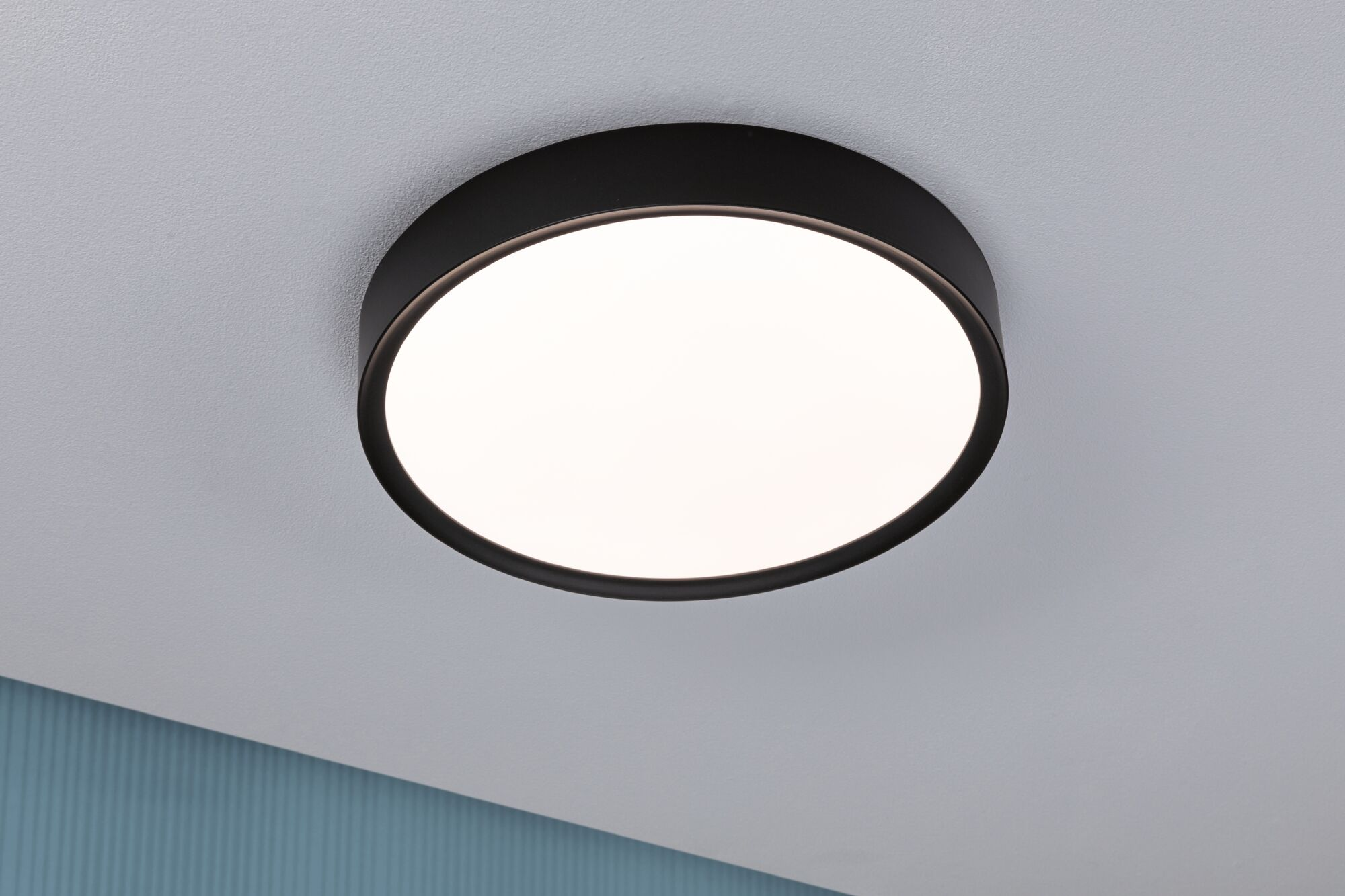Czarna, okrągła lampa sufitowa z białym dyfuzorem jest zamontowana na białym suficie, obok niebieskiej ściany.