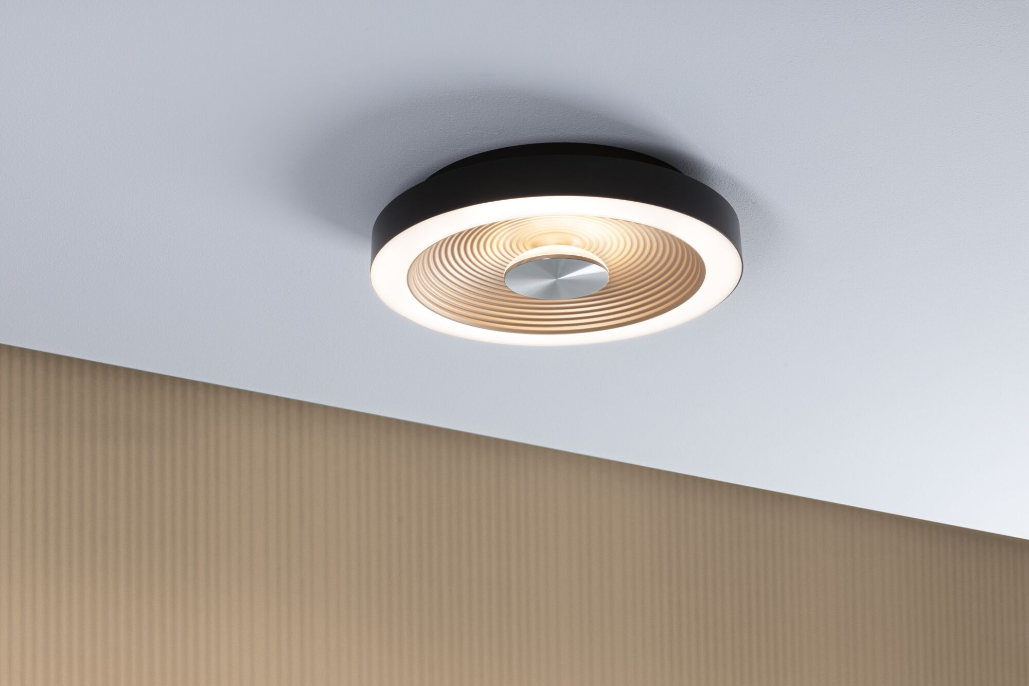 Eine runde, dunkle Deckenleuchte mit silberner Mitte und warm leuchtenden Ringen ist an einer weißen Decke montiert.