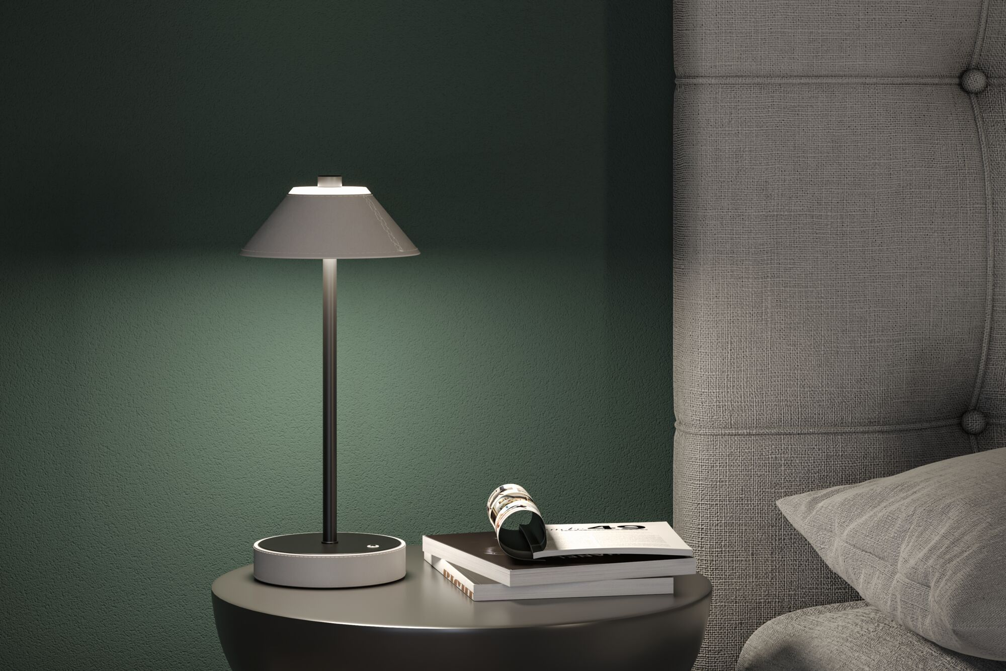 Na stoliku nocnym stoi lampa, książki i okulary obok szarego, pikowanego wezgłowia i zielonej ściany.