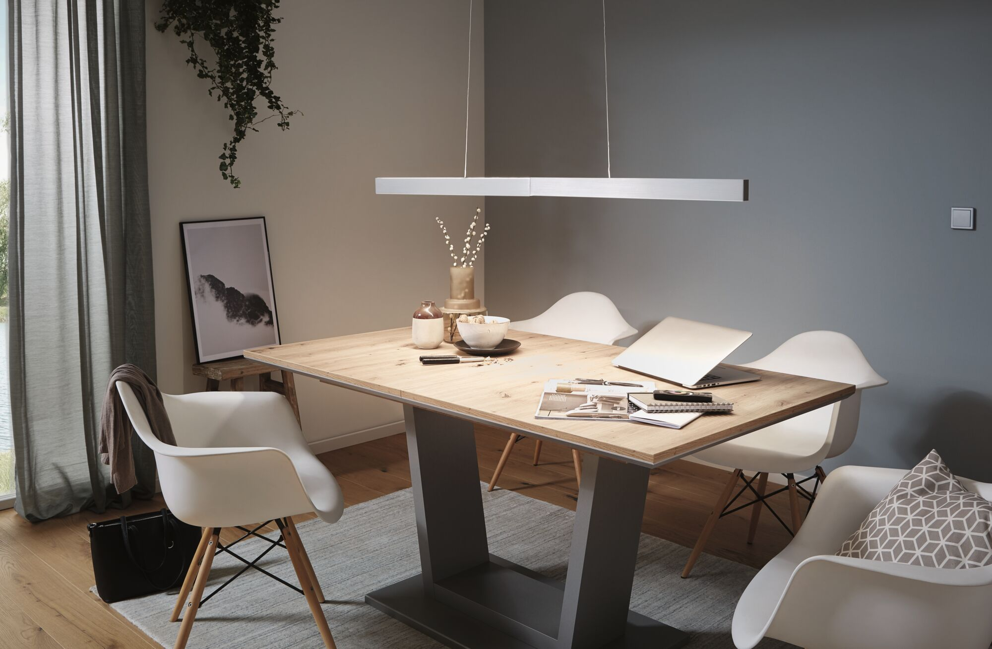 Une salle à manger avec une table en bois, des chaises blanches et un luminaire suspendu. Une image au mur.