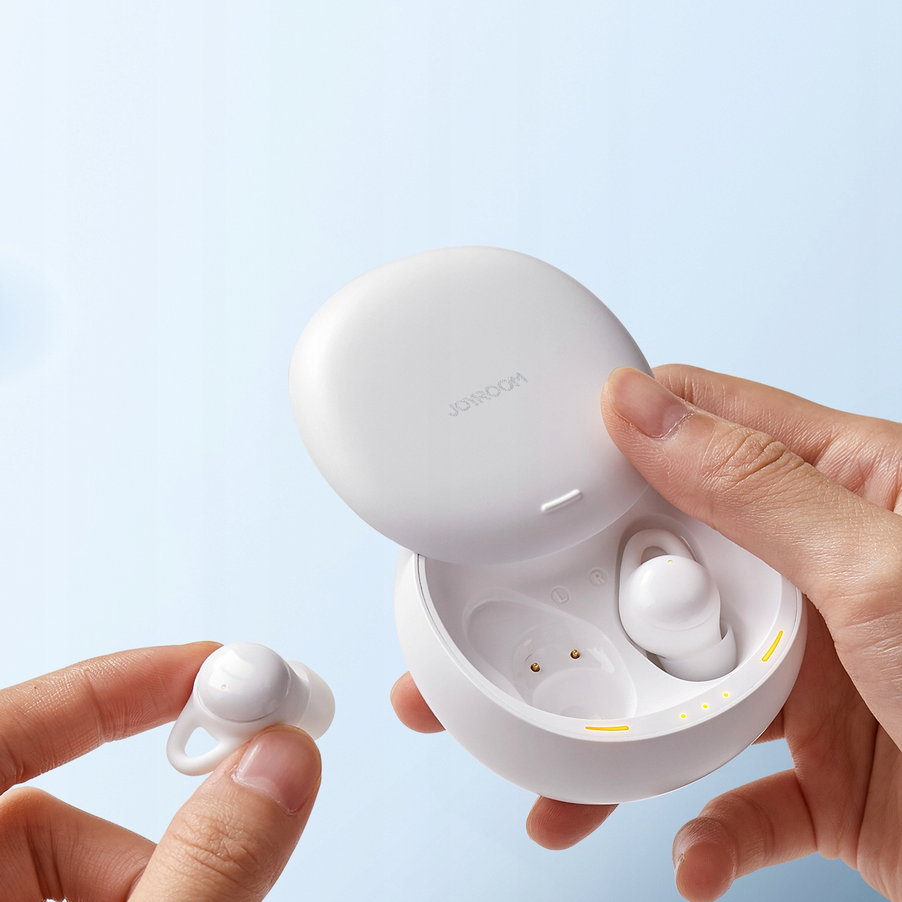 Ręce trzymają białą słuchawkę i jej etui ładujące. Etui jest otwarte, odsłaniając słuchawki.