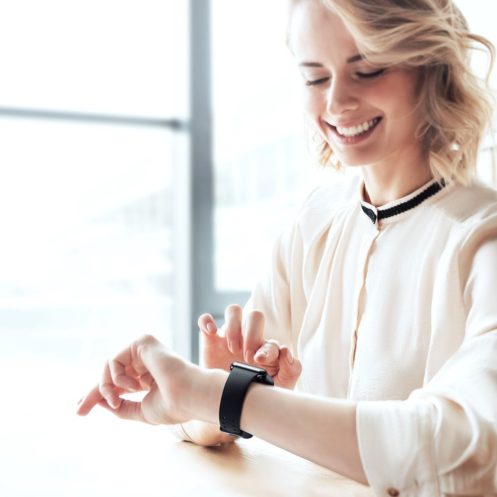 Uśmiechnięta kobieta z czarnym smartwatchem, ubrana w białą bluzkę i siedząca przy stole.