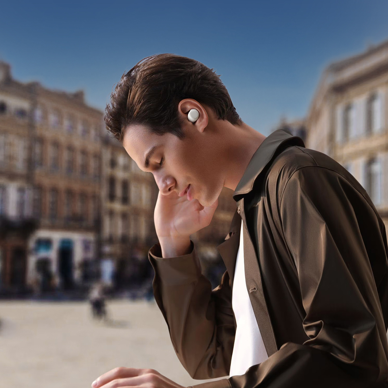 Mężczyzna ze słuchawką w uchu, ubrany w brązową kurtkę na białej koszuli, z rozmytym tłem miasta.