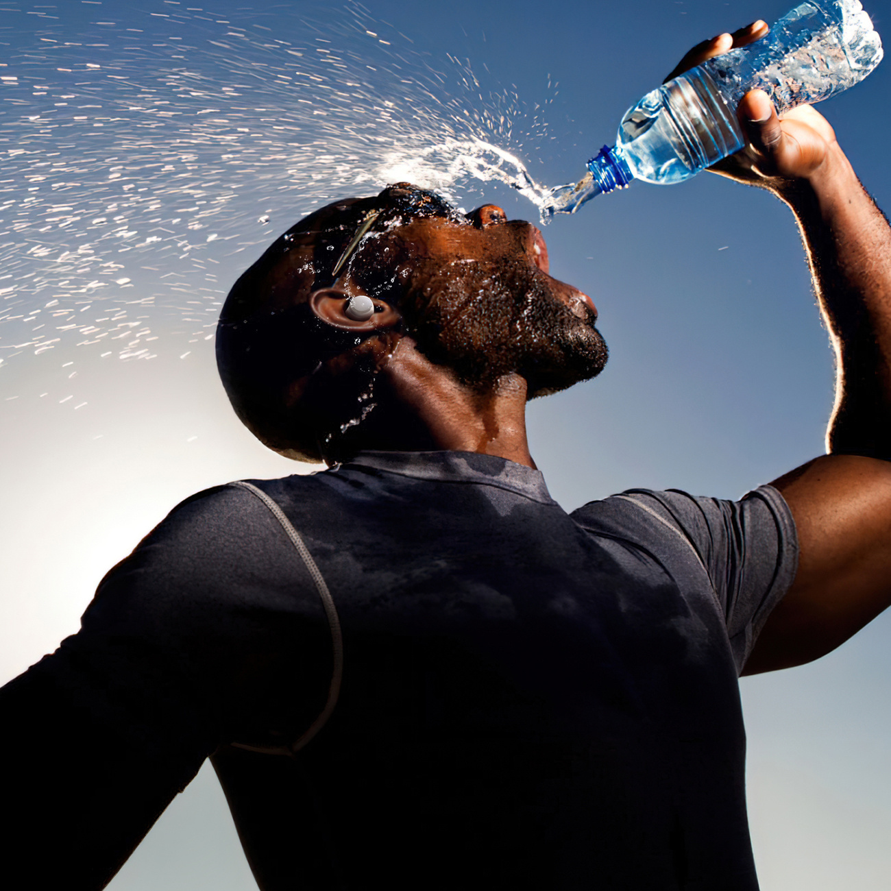 Czarny mężczyzna polewa twarz wodą, ze słuchawką w uchu, na tle jasnego nieba.