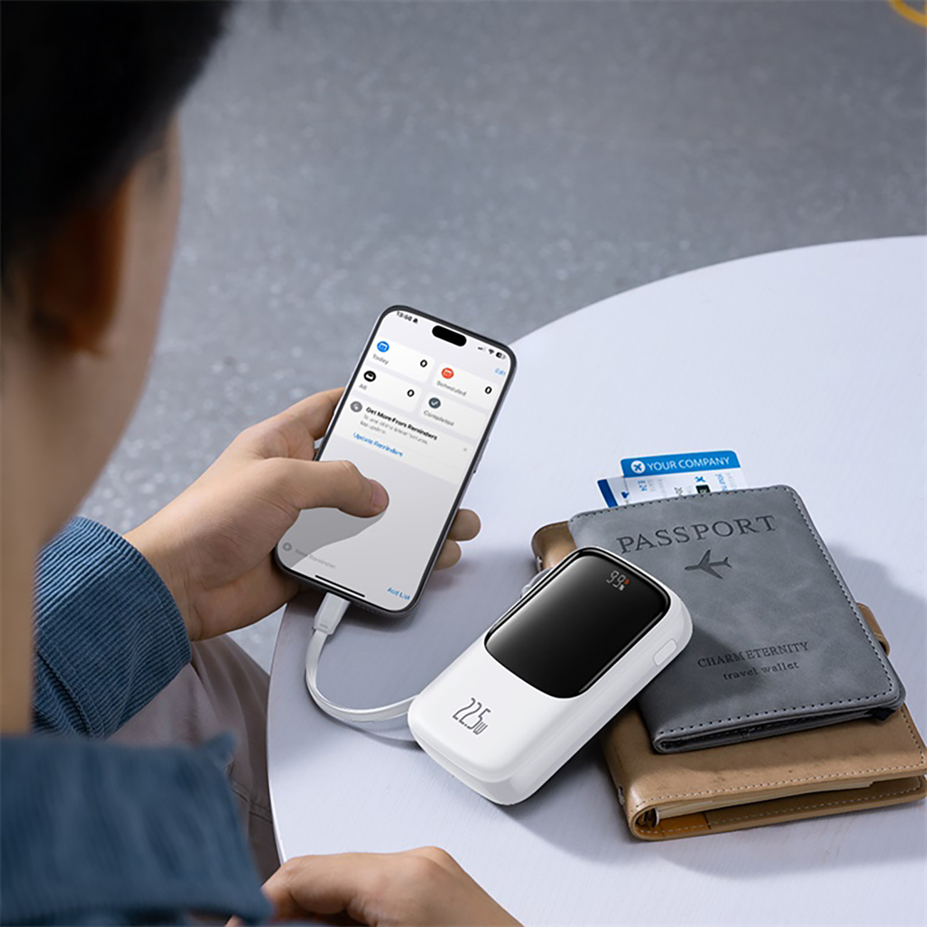 Osoba korzysta ze smartfona i ładuje biały power bank obok paszportu i portfela na białym stole.