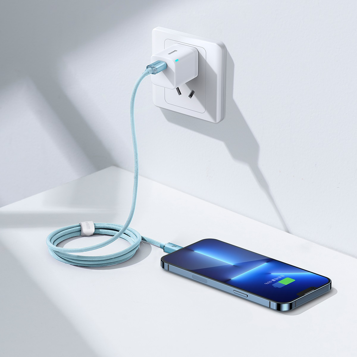 Jasnoniebieski kabel Baseus łączy telefon z ładowarką ścienną, pokazując stan naładowania.