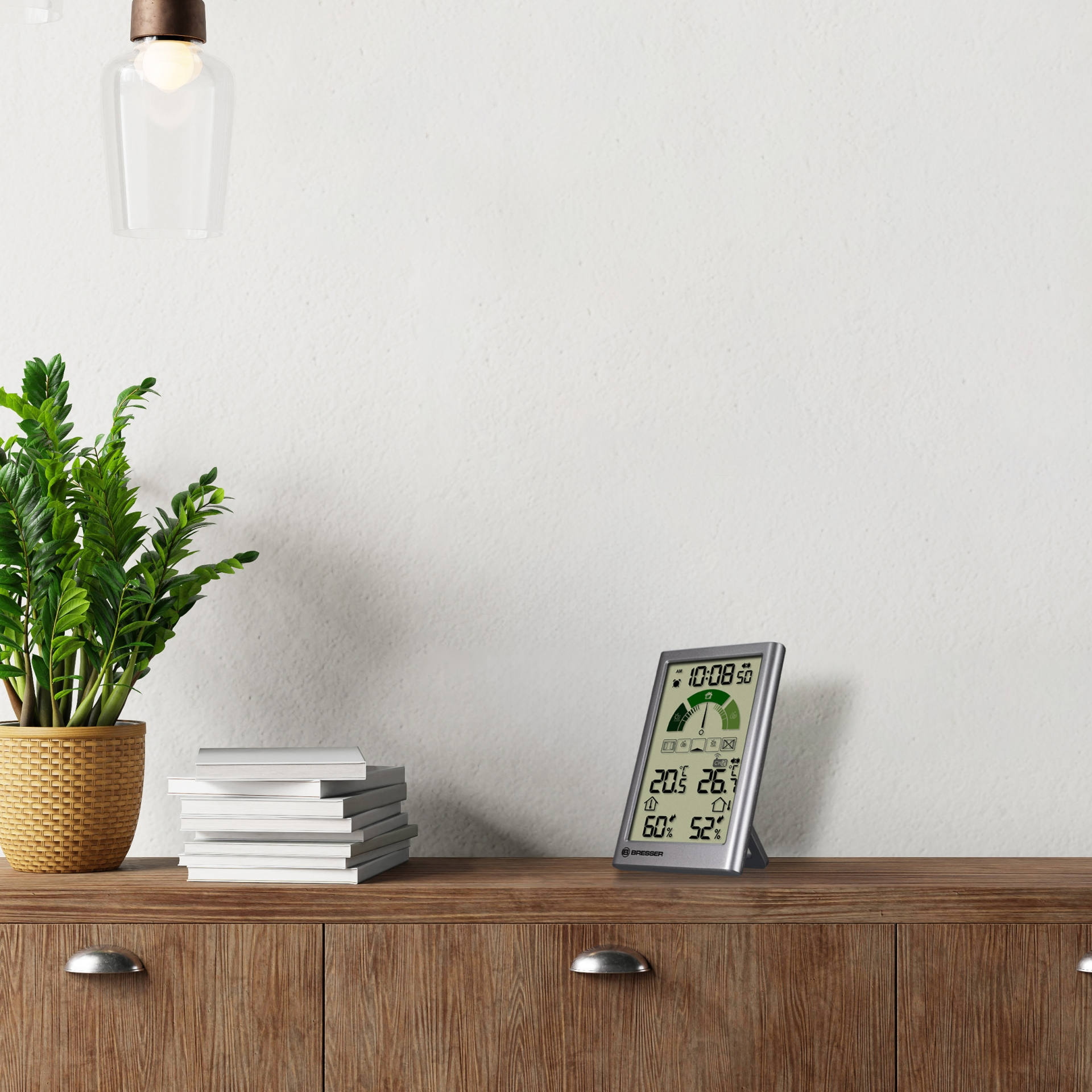 Eine Bresser-Wetterstation auf einem Holzschrank neben einer Pflanze und Büchern zeigt Temperatur und Luftfeuchtigkeit an.