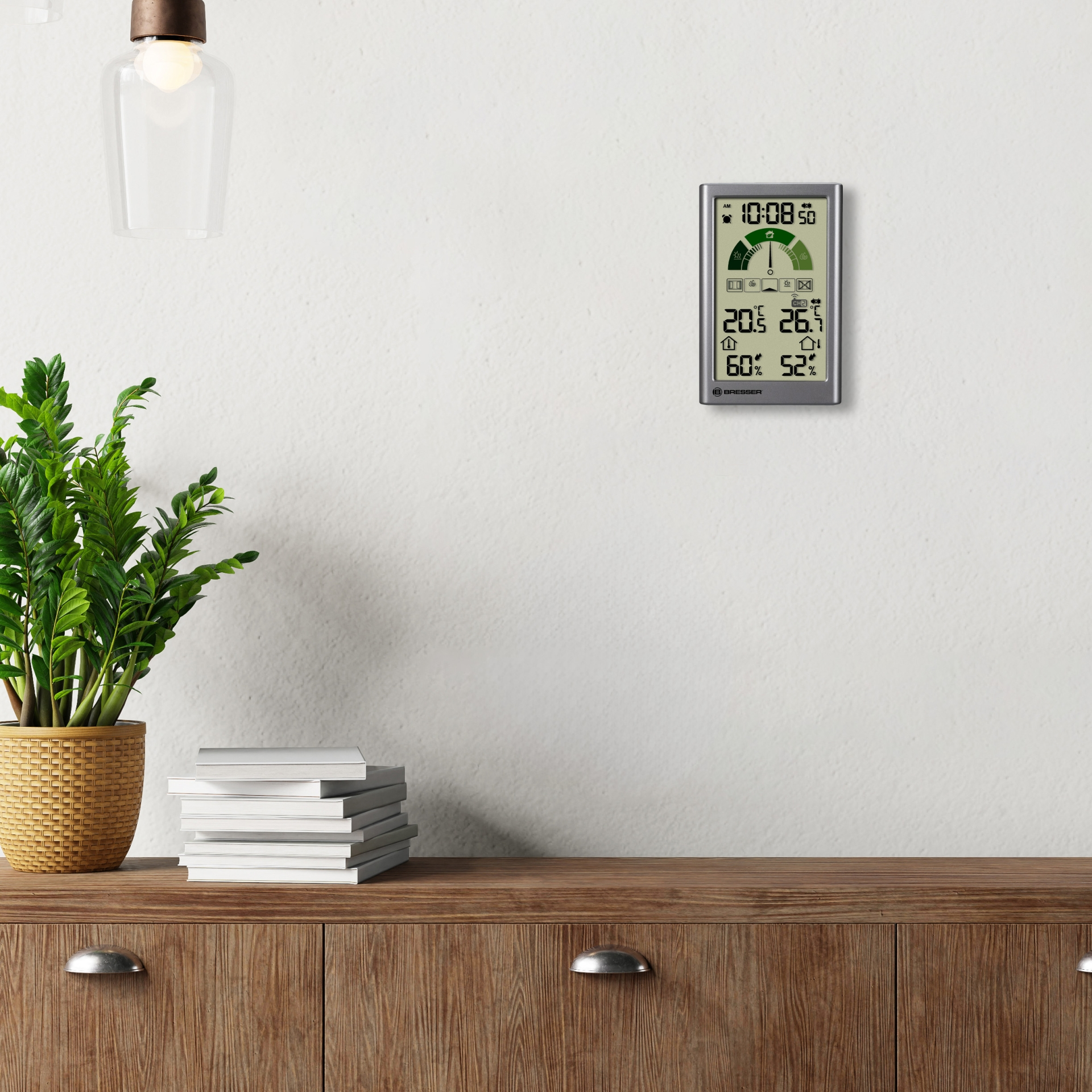 Eine Bresser-Wetterstation auf einem Holzschrank neben einer Pflanze und Büchern zeigt Temperatur und Luftfeuchtigkeit an.
