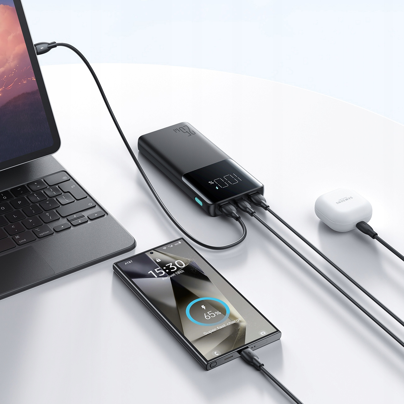 Obraz przedstawia power bank ładujący laptop, telefon i słuchawki na białym stole.