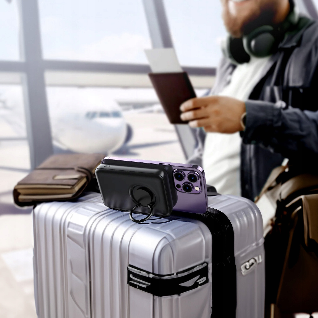 Srebrna walizka z telefonem i ładowarką na górze. Mężczyzna trzyma paszport w tle.