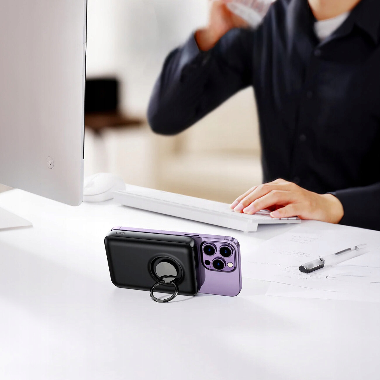 Telefon z czarną ładowarką i stojakiem na pierścień na białym biurku. Osoba pracuje w pobliżu.