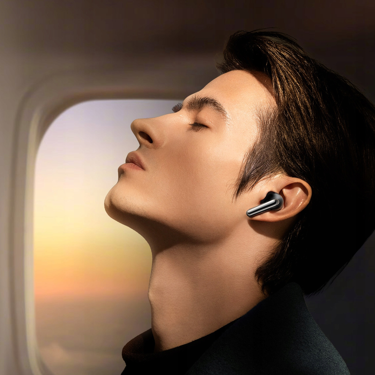 Un uomo con un auricolare nell'orecchio, che guarda in alto. È su un aereo vicino a un finestrino con vista sul tramonto.