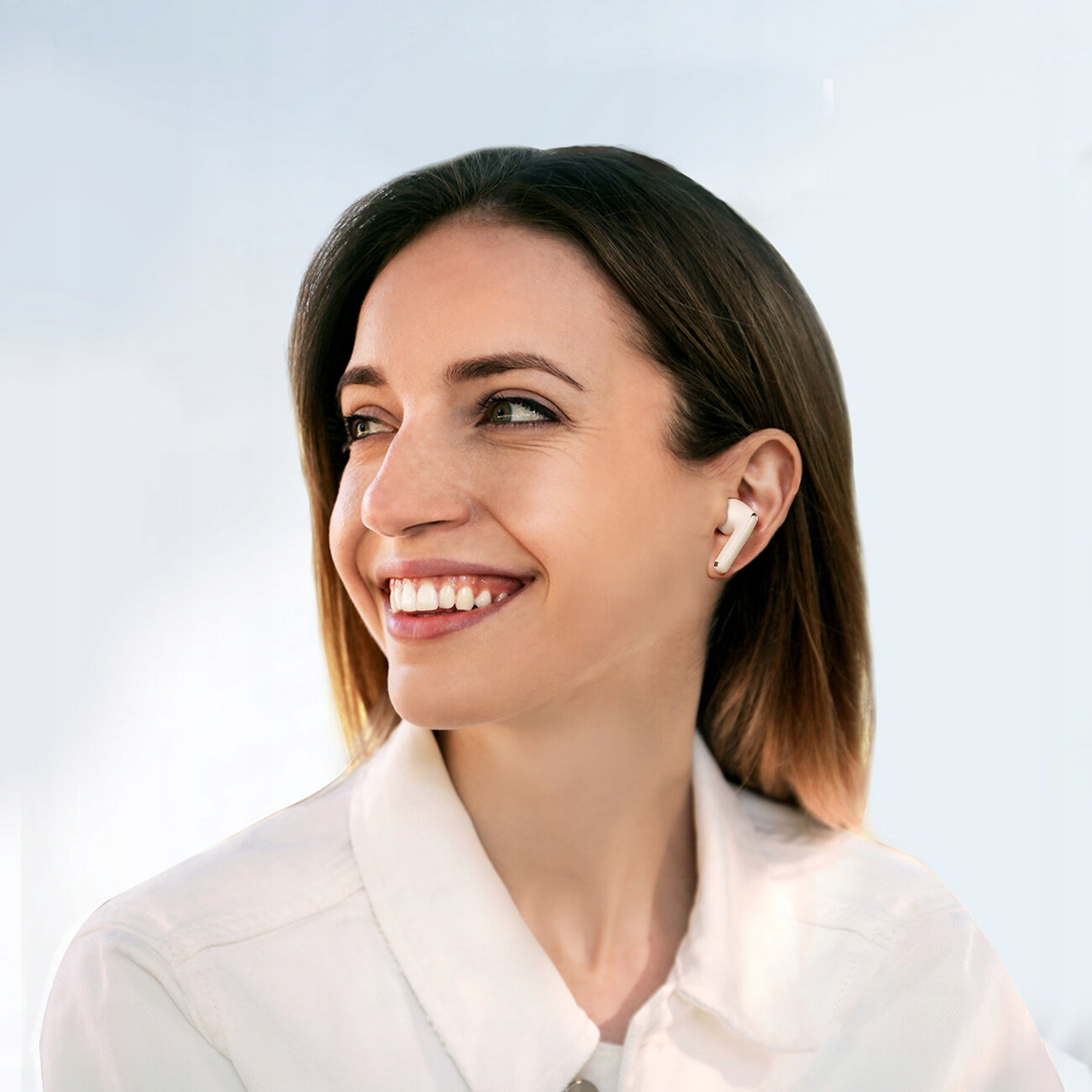Uśmiechnięta kobieta, nosząca białe bezprzewodowe słuchawki i białą koszulę, na tle jasnego nieba.