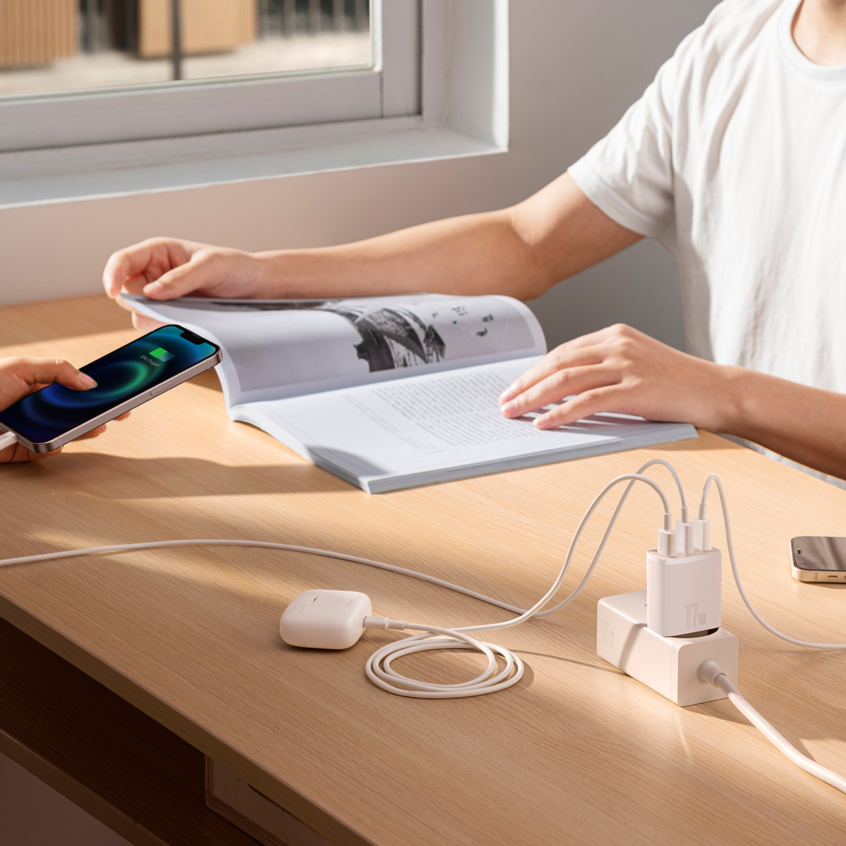 Osoba czyta książkę, podczas gdy telefon i słuchawki są ładowane za pomocą ładowarki 17W na drewnianym stole.