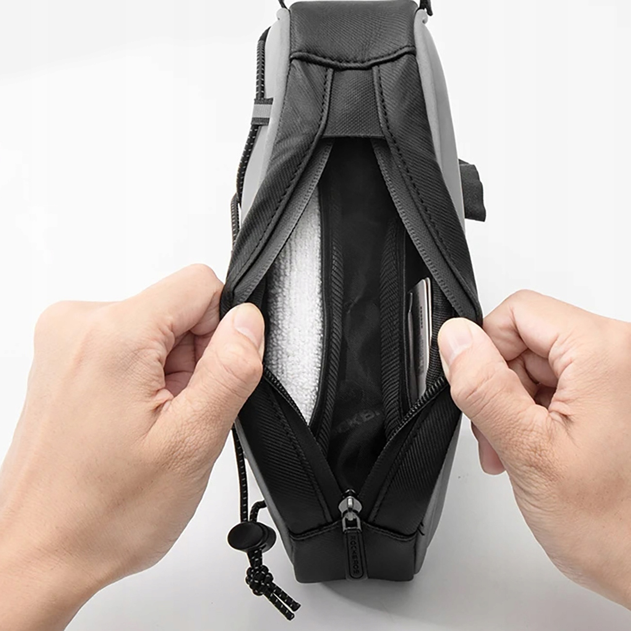 Ręce otwierają szarą i czarną torbę rowerową, odsłaniając białą szmatkę i telefon w środku. Widoczny zamek błyskawiczny i sznurek.