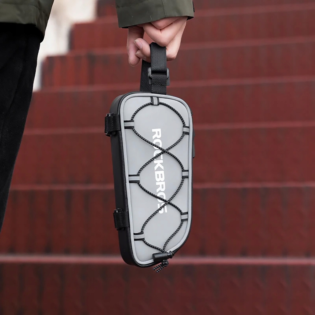 Ręka trzyma szarą torbę rowerową z czarnymi paskami i gumką na tle czerwonych schodów.