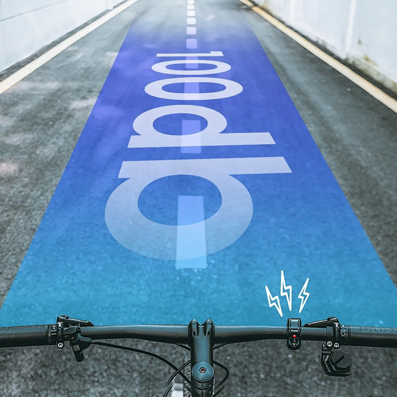 Rower na drodze oznaczonej na niebiesko napisem „100dB”, wskazującym poziom dźwięku, widzianej z perspektywy rowerzysty.
