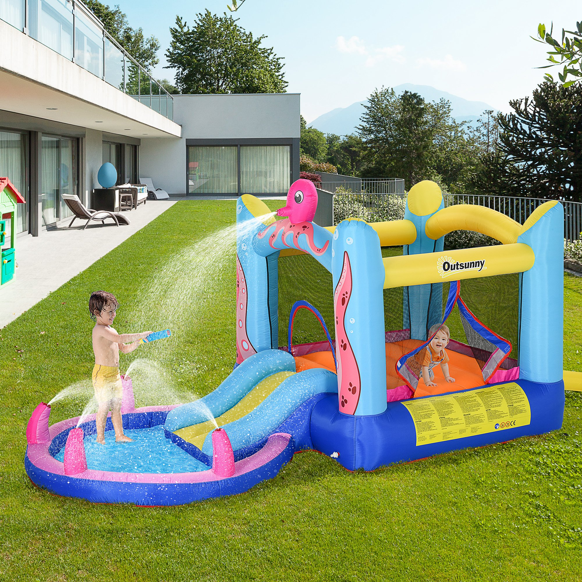 Una colorida casa de rebote inflable y un tobogán acuático se asientan sobre hierba verde con un niño jugando con una pistola de agua.