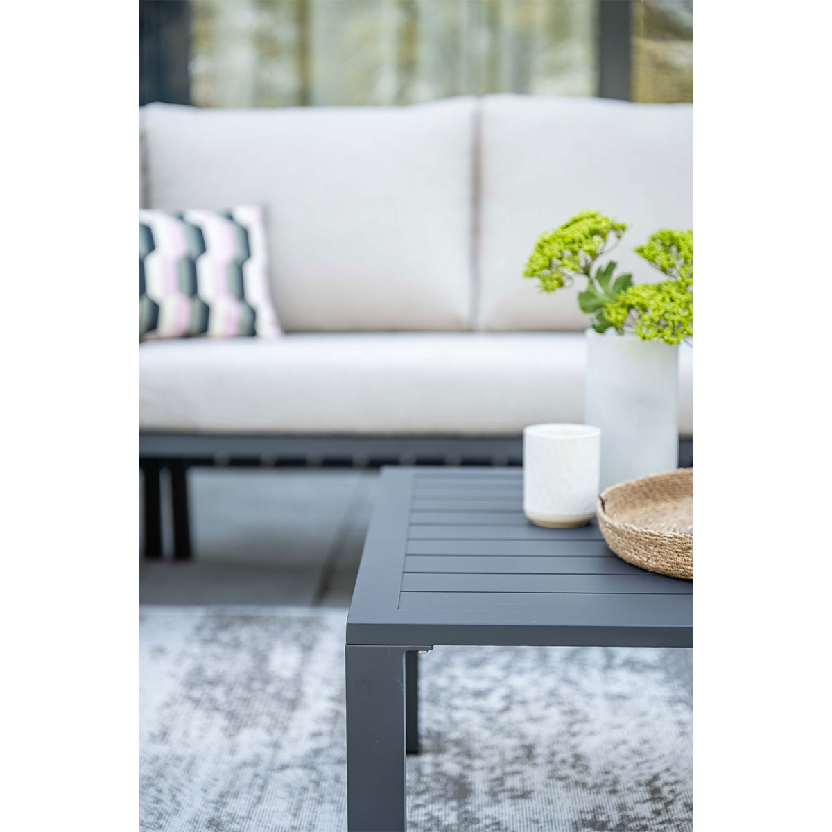 Square gray table with decor, sectional sofa and patterned rug on a patio.