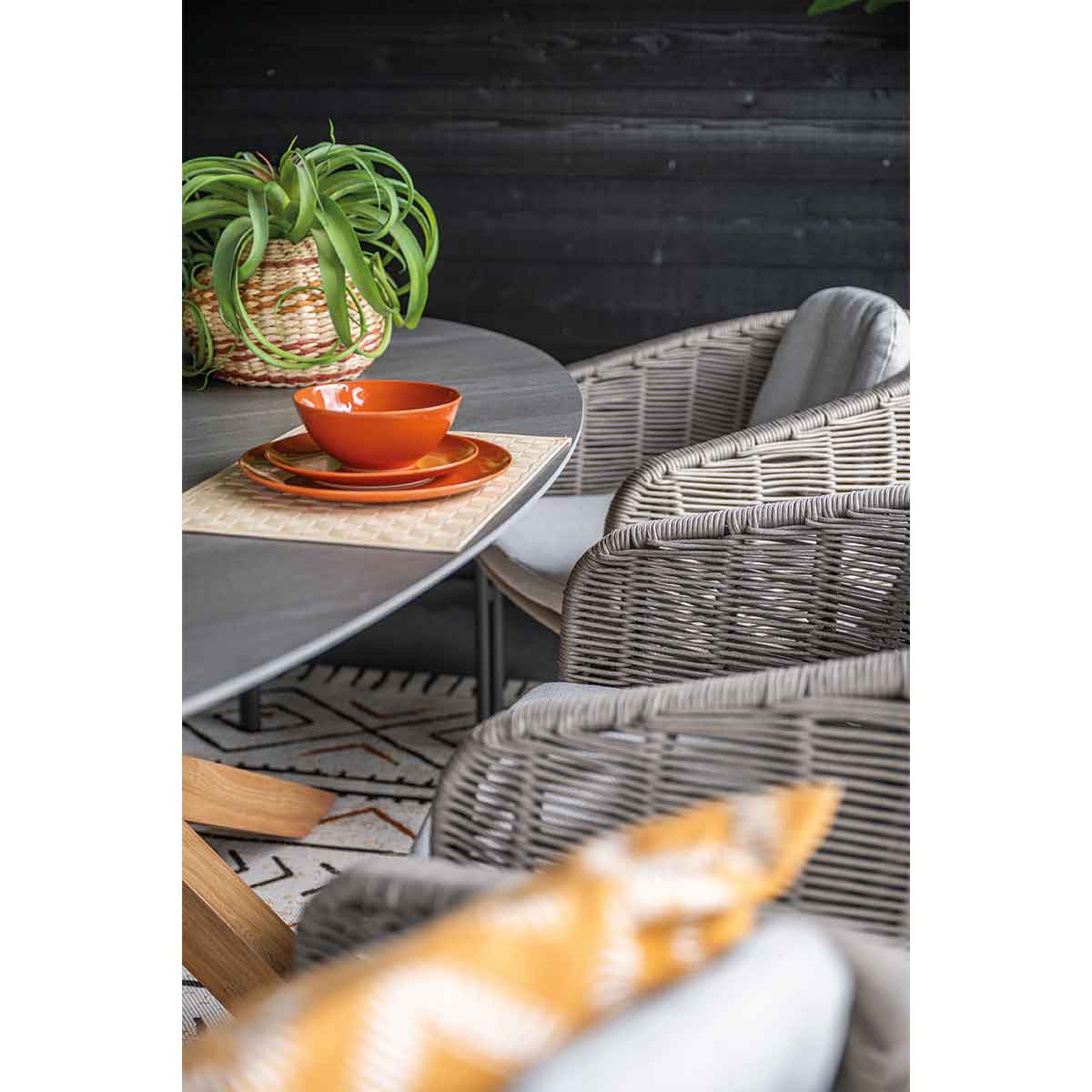 Detail of outdoor table setting with woven chairs, orange dishes, and green plant.