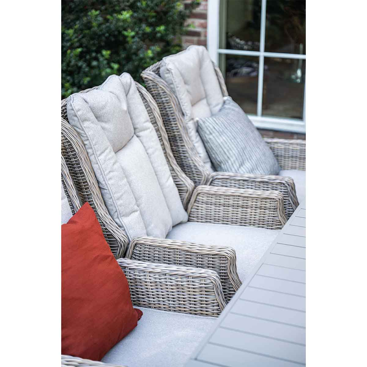 Buiten zithoek met grijze wicker stoelen, kussens en een rood kussen, geplaatst tegen een bakstenen muur en groen.