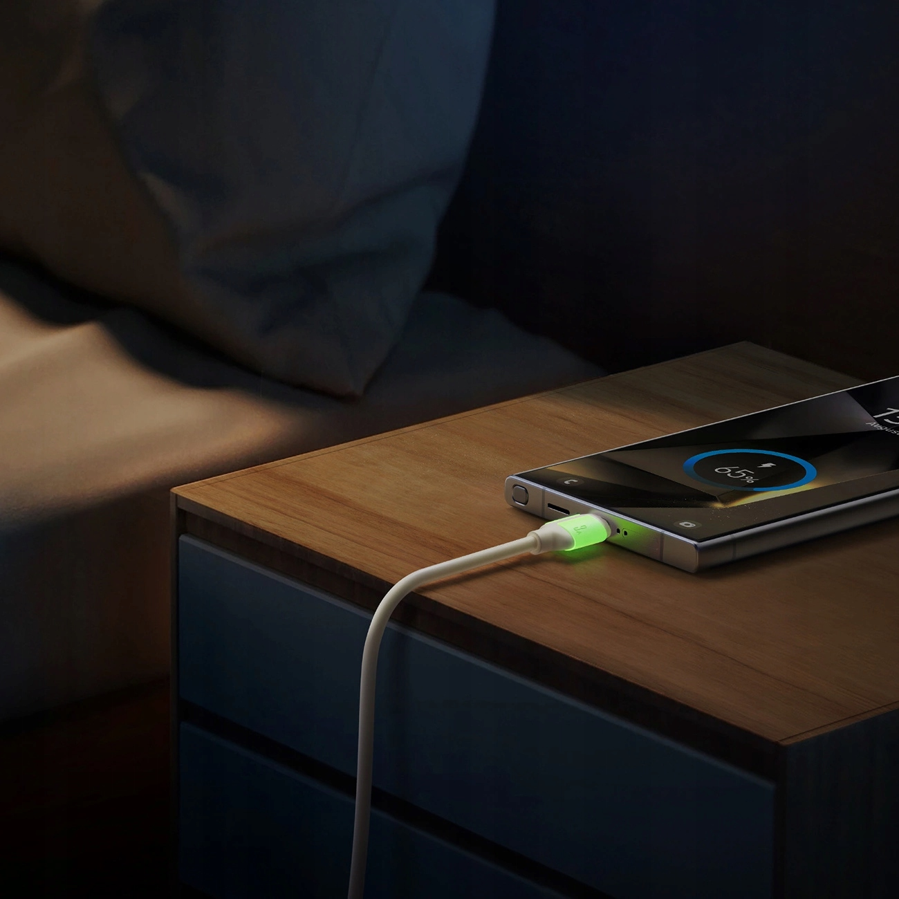 Un smartphone est en charge sur une table de chevet en bois, connecté à un câble blanc avec un connecteur vert brillant.