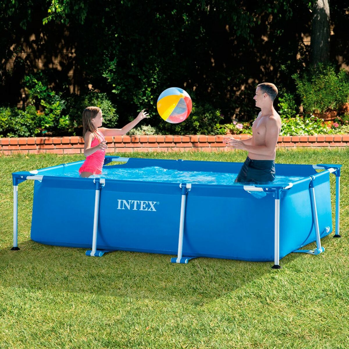 Una niña y un hombre juegan con una pelota de playa en una piscina rectangular azul Intex sobre césped verde bajo árboles.
