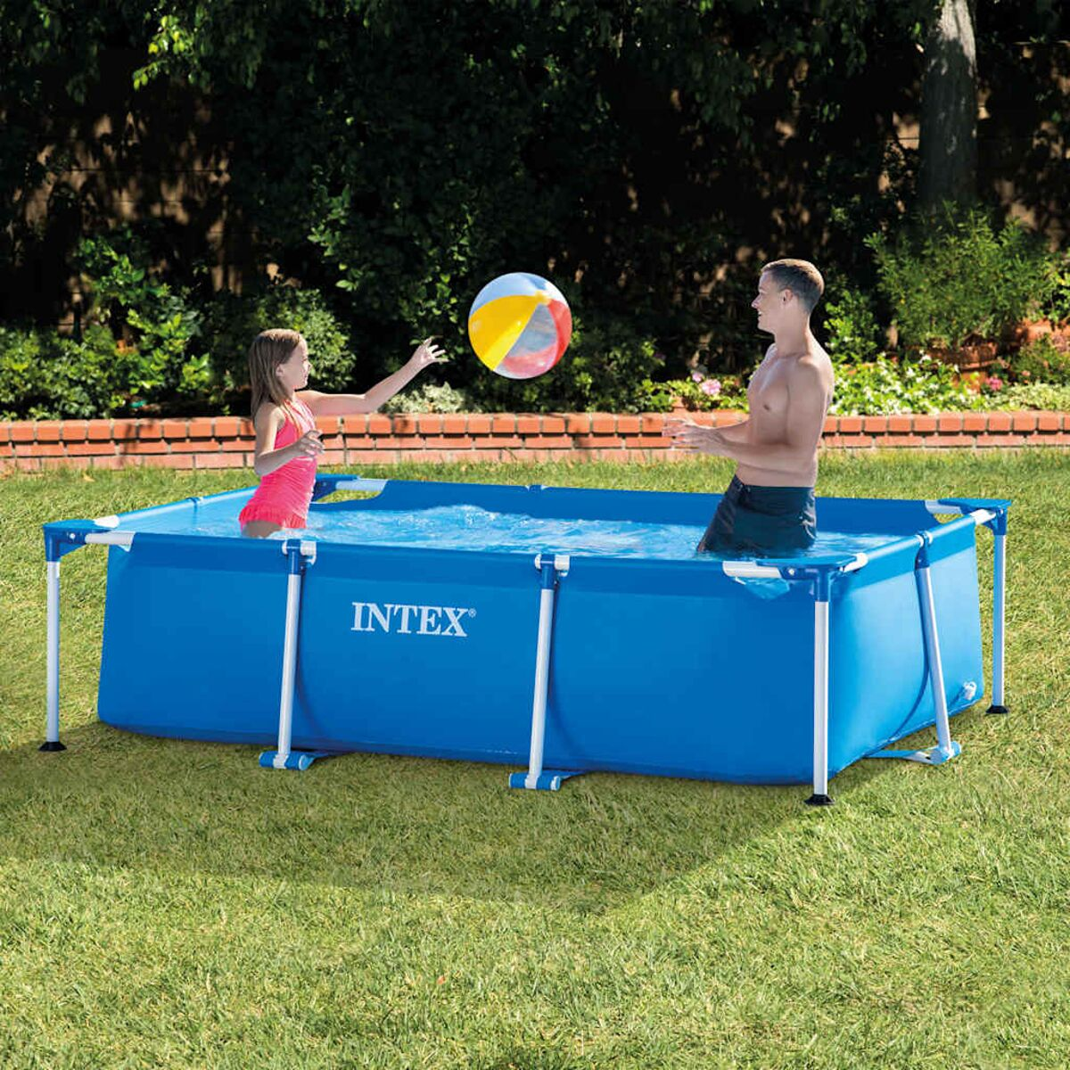 Una niña y un hombre juegan con una pelota de playa en una piscina rectangular azul Intex sobre césped verde bajo árboles.