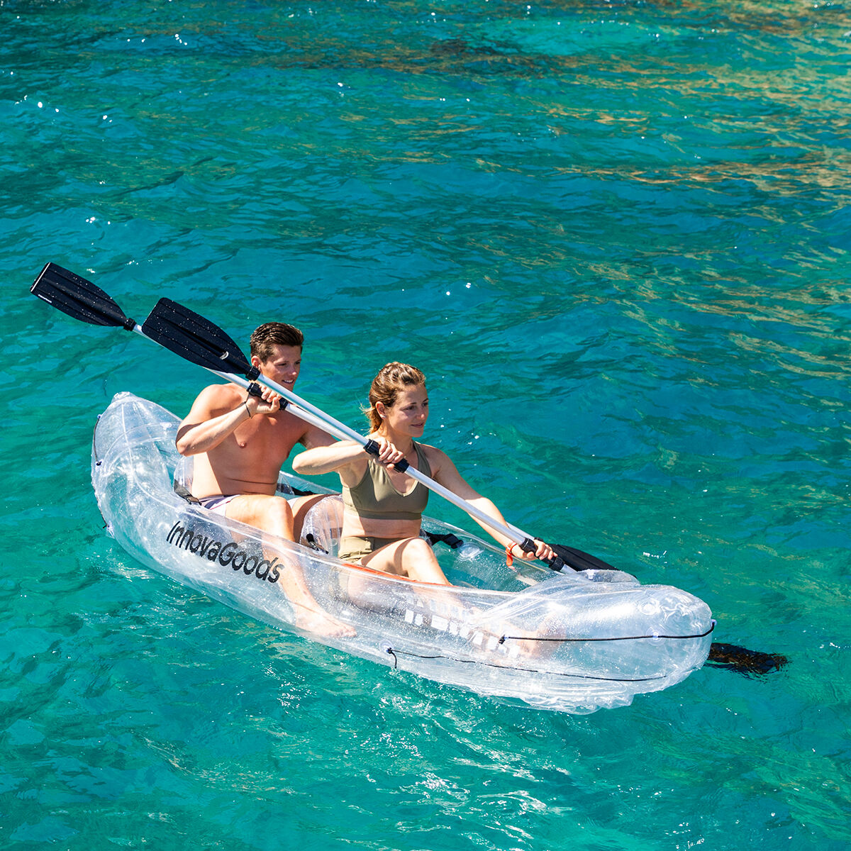 Un hombre y una mujer reman en un kayak transparente en aguas turquesas, disfrutando de un día soleado en el mar.