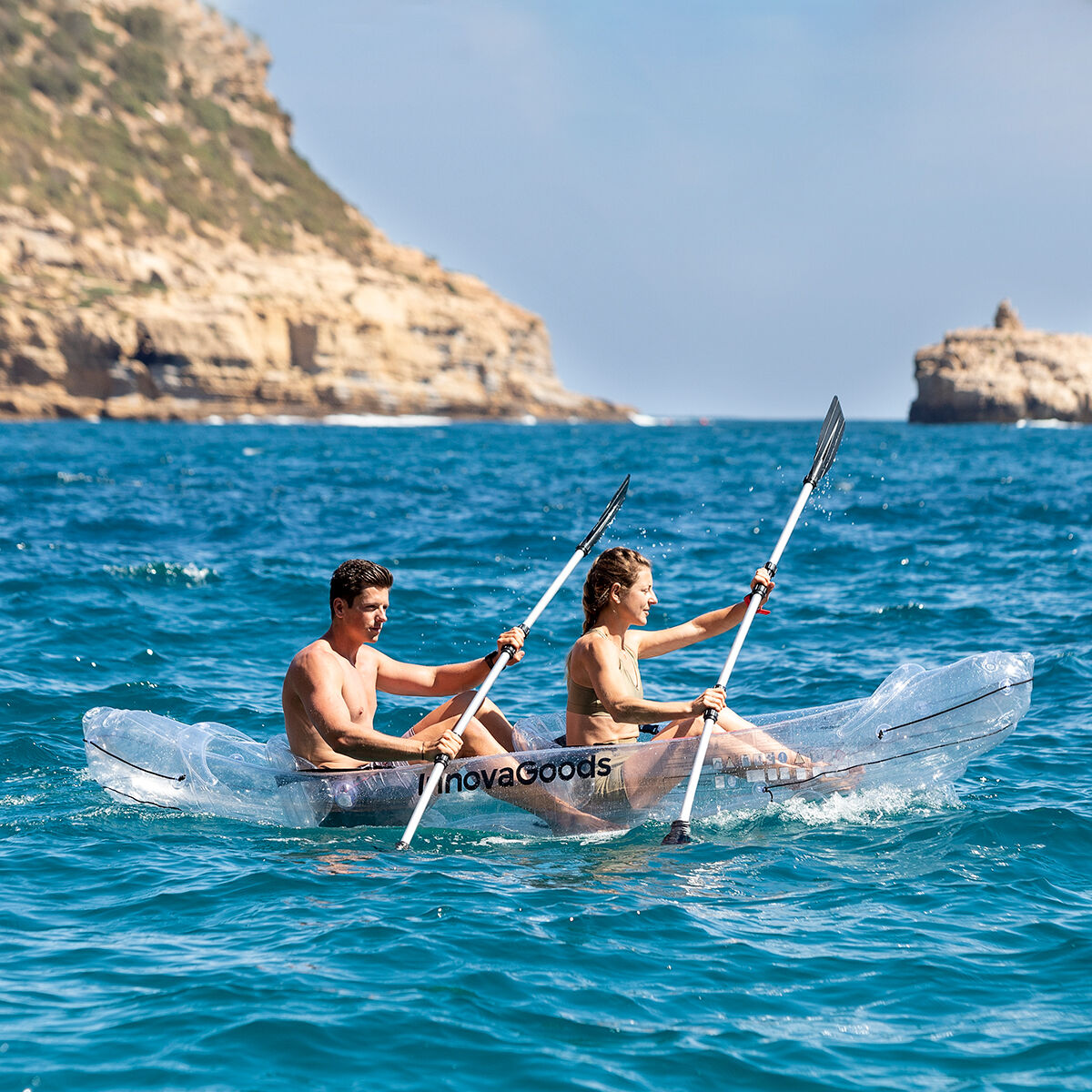 Un hombre y una mujer reman en un kayak transparente en el océano, con acantilados visibles al fondo bajo un cielo azul.