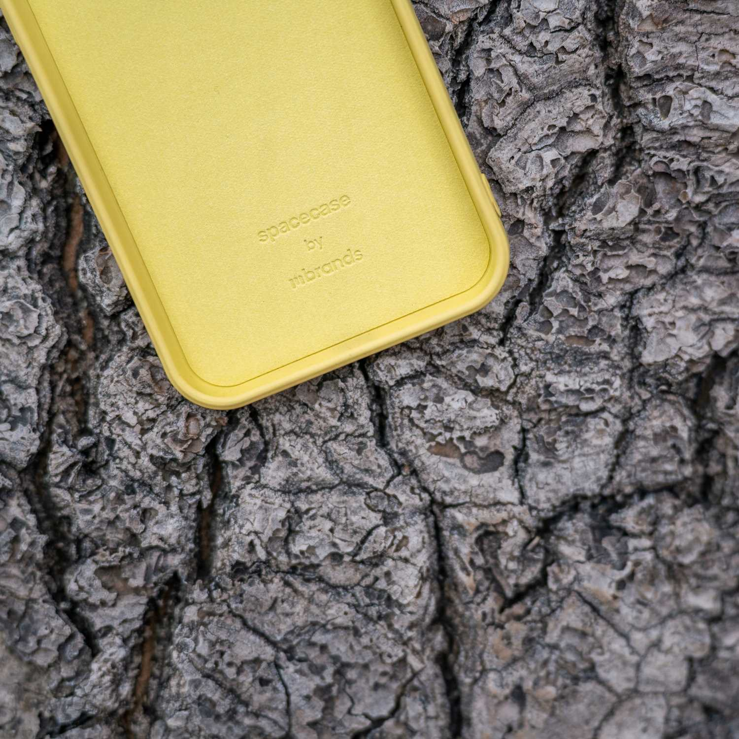 Żółte etui na telefon z tekstem „spacecase by mbrands”. Tło z szorstkiej, szarej kory drzewa.