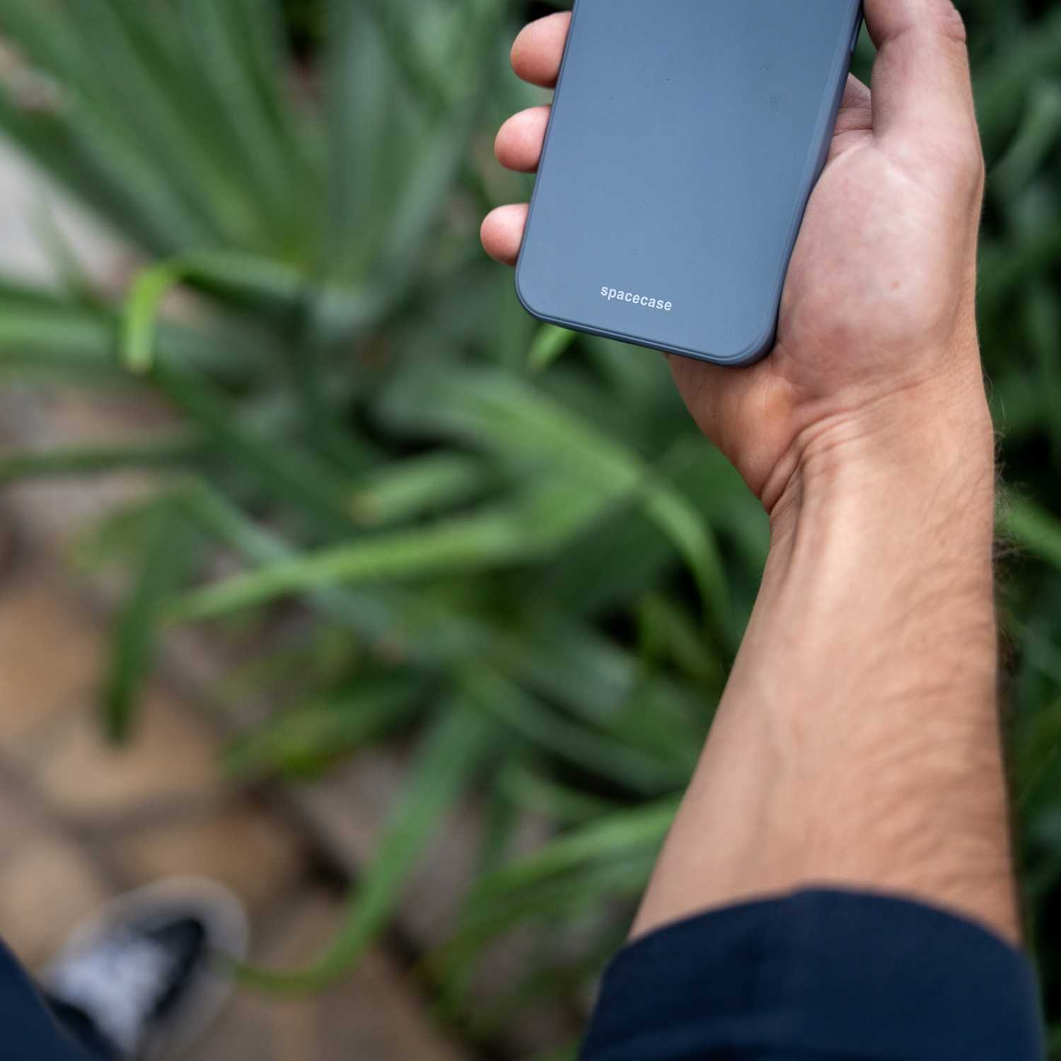 Ręka trzyma niebieskie etui na telefon, z zielenią i rozmytą kamienną ścieżką w tle.