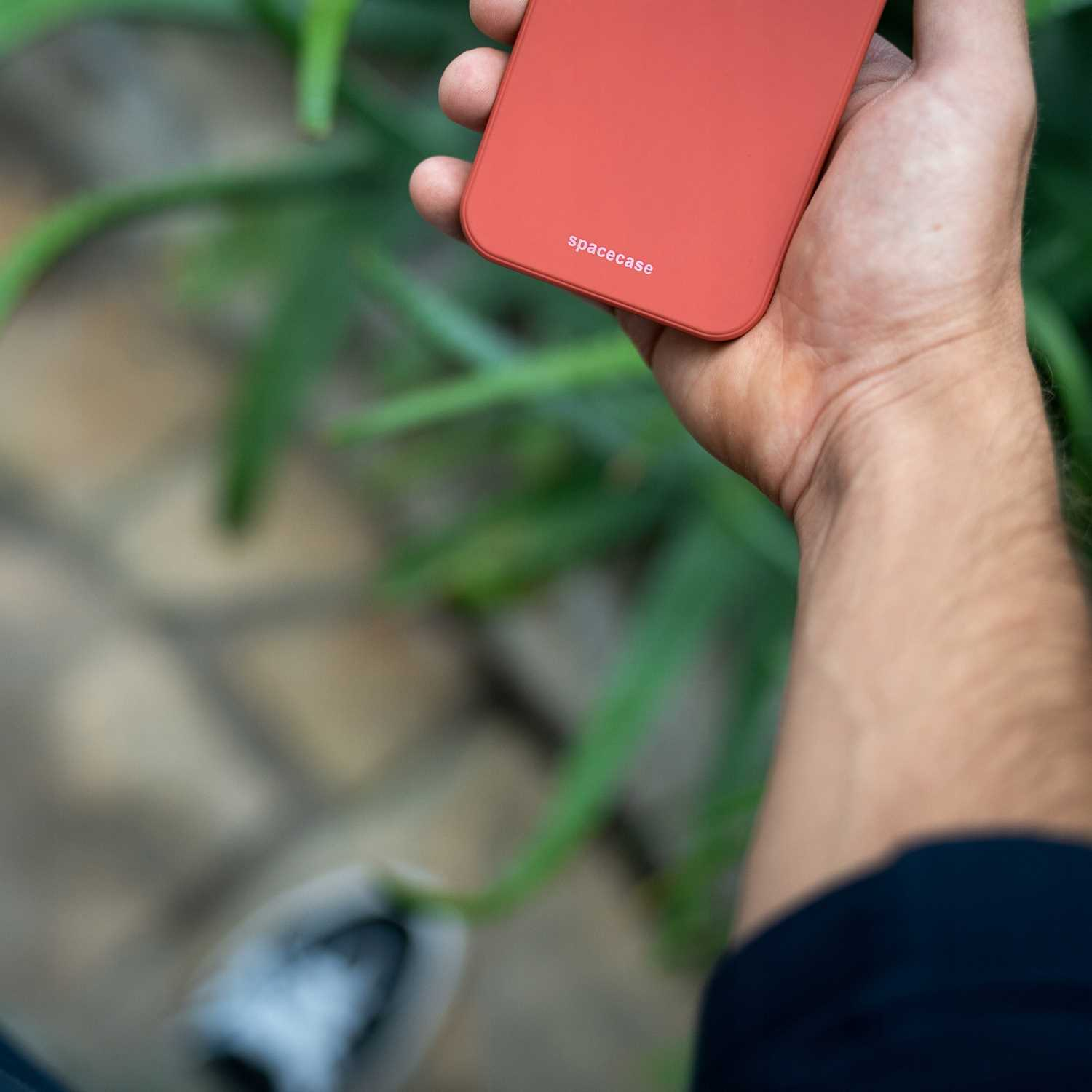 Czerwone etui na telefon z napisem „spacecase” trzymane nad rozmytymi liśćmi i ziemią.