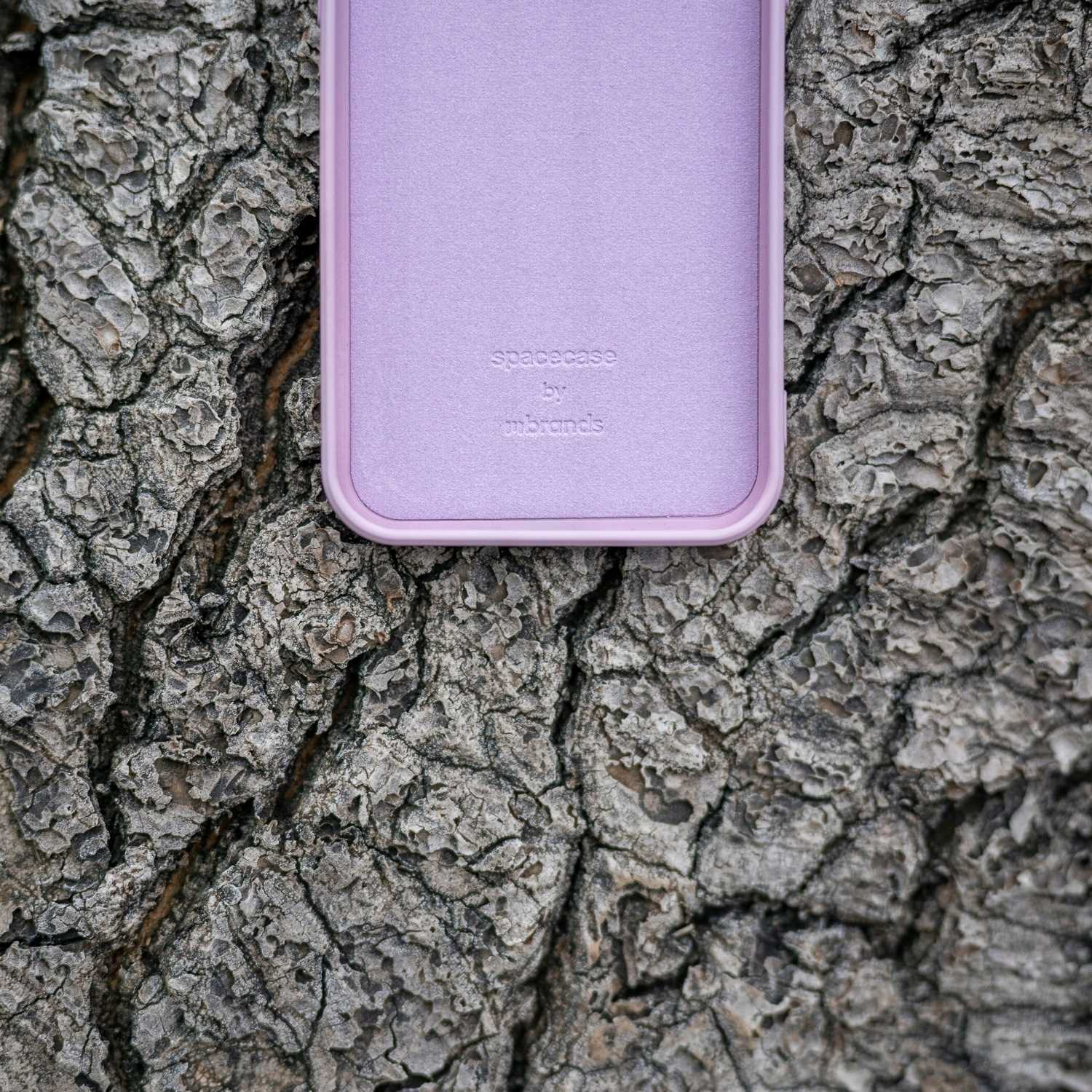 Liliowe etui na telefon leży na korze drzewa, z nazwą marki. Kora ma szorstką, teksturowaną powierzchnię.