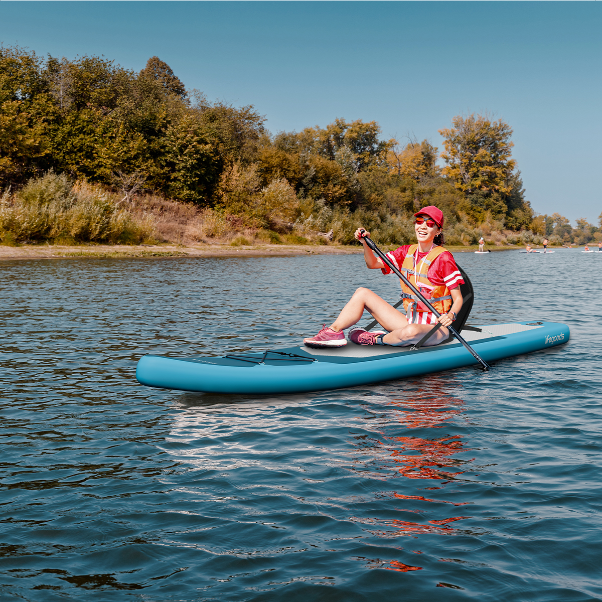 Een vrouw peddelt op een blauw paddleboard in een meer, omringd door bomen onder een heldere hemel.