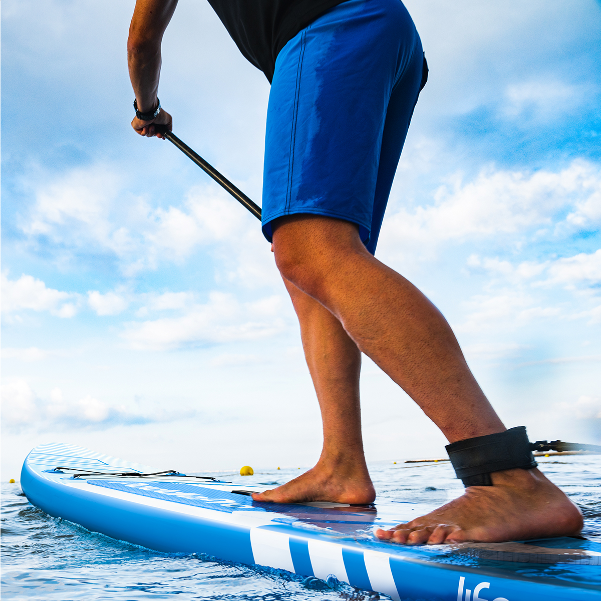 Person mit blauen Shorts, die auf einem Paddleboard auf dem Wasser unter einem bewölkten Himmel steht.
