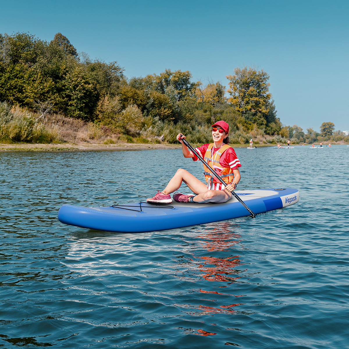 Vrouw peddelt op een blauwe paddleboard op het water met bomen en andere paddleboarders op de achtergrond.