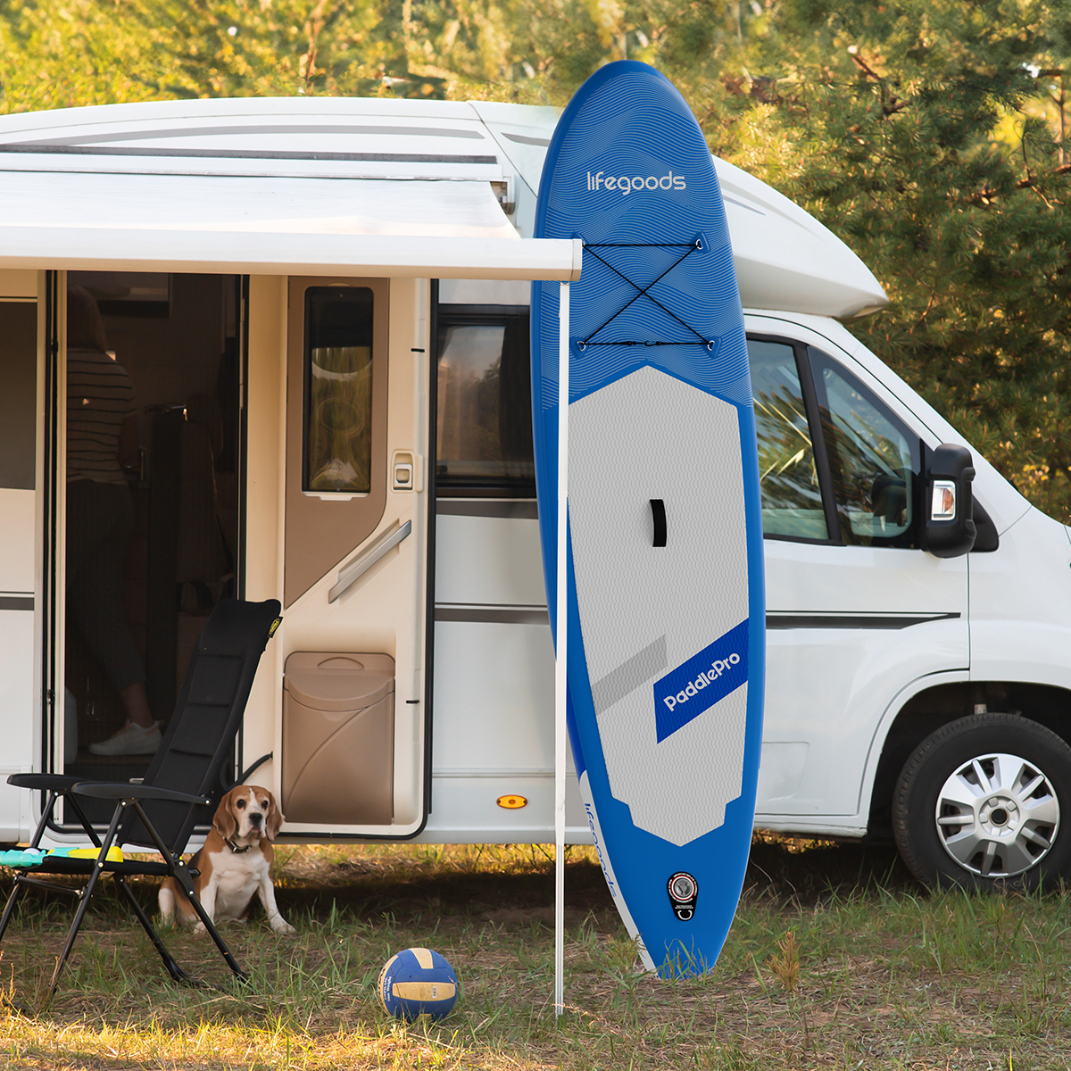 Een witte camper met een blauw paddleboard, een zwarte stoel, een hond en een volleybal in een grasveld.